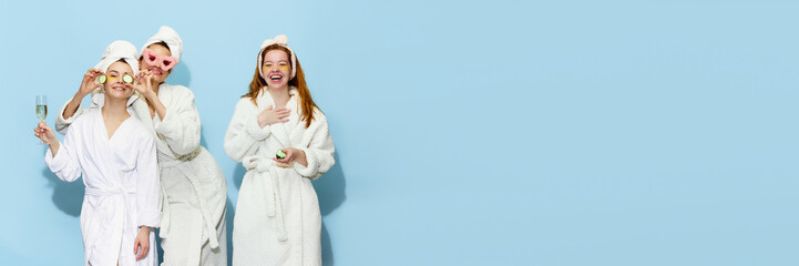Hen party. Happy, smiling, beautiful girls in bathrobes getting ready, taking care after skin, having fun over blue studio background. Concept of youth, beauty, friendship. Copy space for ad. Banner