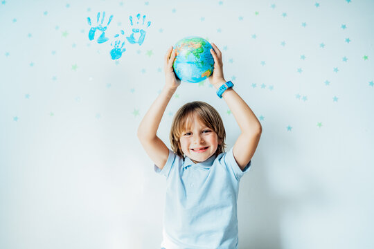 Smiling caucasian boy holding an earth globe model over his head on the background with stars and family hand prints on the white wall. Save the planet, Earth Day. Global peace and unity concept. - Powered by Adobe
