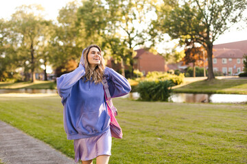Attractive curly blonde woman walk on the city park street. Girl wear purple hoodie, pink bag and look happy and smiles. Woman put on hood while go on the street, look at side.