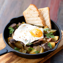 Fried potatoes with egg, bread and meat in frying pan. Rustic lunch on wooden cutting board. Homemade food. Hearty high-calorie lunch. Side view. Soft focus. 