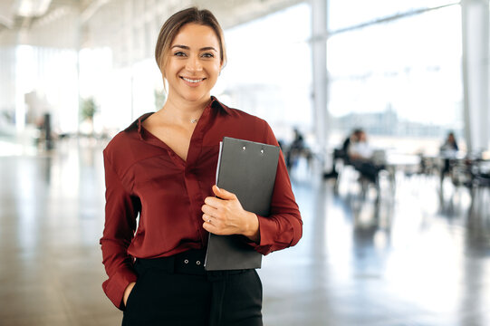 Portrait of a gorgeous successful confident positive blonde caucasian business lady dressed in an elegant wear, stands in a business center, holds documents, looks at camera, smiling friendly