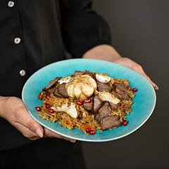 Plate of pilaf with lamb. Pilaf with meat is decorated with pomegranate seeds and head of garlic on blue plate in hands of woman. Traditional oriental dish. Middle Asian cuisine. Black background. 