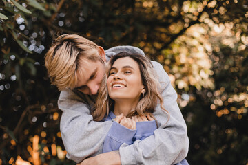 Fototapeta premium Portrait of beautiful blonde curly girl looking away with happy smile while man kissing her neck. Happy couple walk in park.