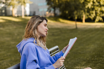 Young student woman holding personal diary sitting on bench in summer park, look happy and smiling....