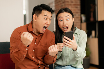 Emotional asian couple celebrating success, looking at cellphone screen and emotionally reacting to...