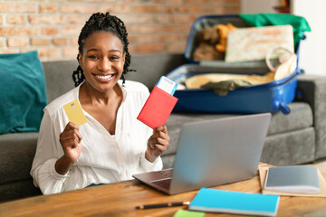 Happy black woman using laptop, buying travel tickets, holding passport and credit card, sitting with unpacked suitcase