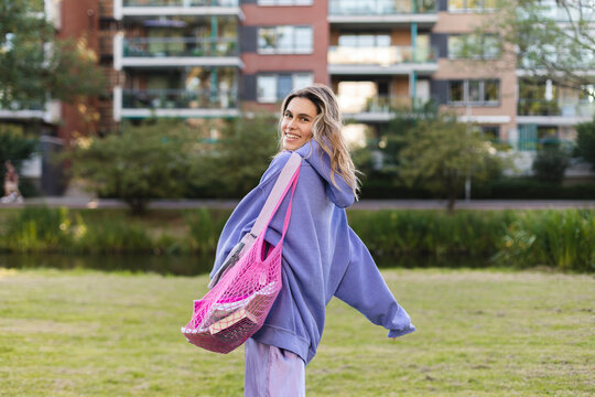 Pretty young caucasian woman with curly blonde hair turns around walking in the park. Blonde smiling, dressed in casual purple hoody and skirt. Rest and recovery concept. Happy girl hold pink tote bag