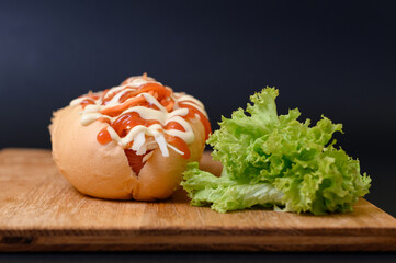 Hot dog covered with ketchup and mayonnaise on a wooden board on a black background.