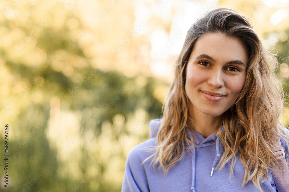 Wall mural Close up portrait attractive young happy woman with fresh and clean skin stands outside in the green park. Smiling curly blonde wear purple hoody and look at camera. Lifestyle, female beauty concept.