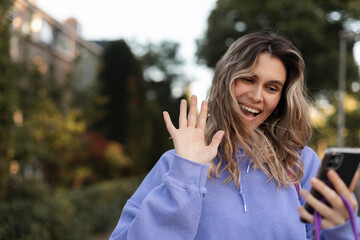 Happy young blonde curly woman standing on city street, outdoors holding phone looking at screen waving hand video calling distance friend online in mobile app using smartphone video chat application.