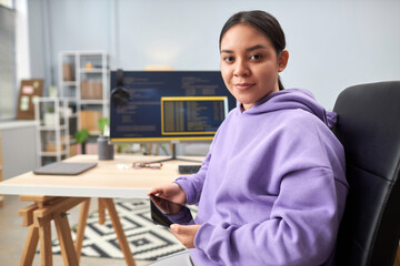 Portrait of young woman as IT programmer looking at camera at workplace with code screens in background, copy space