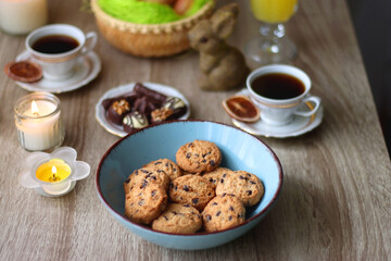 Easter eggs in the basket, bowl of cookies, chocolate pralines, Easter bunny figurine, cups of tea, glasses of juice, flowers and lit candles on the table. Selective focus.