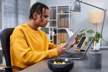 Side view portrait of young black man working or studying at home office workplace and holding documents