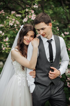 Happy young couple standing near beautiful flowers. The bride gently hugged her groom behind the shoulders. The young embrace tenderly, looking down. Wedding portrait. Spring wedding