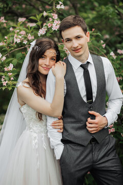 Happy young couple standing near beautiful flowers. The bride gently hugged her groom behind the shoulders. Young people are gently hugging, looking at the camera. Wedding portrait. Spring wedding