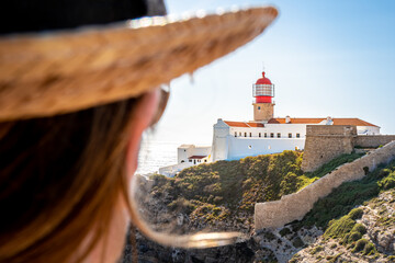 A unrecognizable female tourist with hat gazes at the Farol do Cabo de São Vicente lighthouse atop...