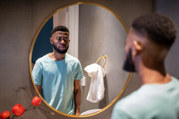 Unhappy upset 30s African American man looking in mirror, standing in bathroom at home, sad...
