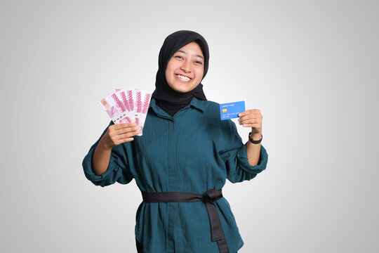 Portrait of cheerful Asian muslim woman with hijab, showing one hundred thousand rupiah while holding a credit card. Financial and savings concept. Isolated image on white background