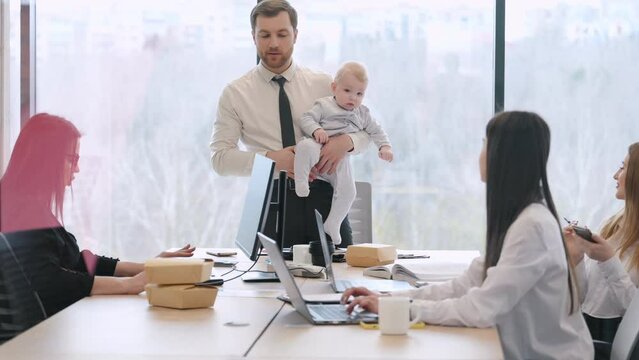 Man is standing, talking to empoyees and holding little baby boy in hands. Group of people working in the modern office together.