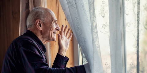 Smiling senior man wearing simple shirt waving with hand through window glass covering with white...