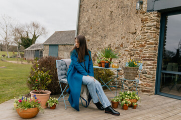 Fototapeta na wymiar Positive young caucasian girl is looking down with smile resting on summer terrace with flowers on background old house. Brunette wears jeans and blue coat. Technology concept, lifestyle 