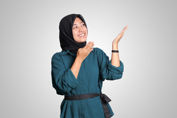 Portrait of excited Asian hijab woman in casual outfit standing against white background, showing product and pointing with hand and finger to the side. Advertising concept.