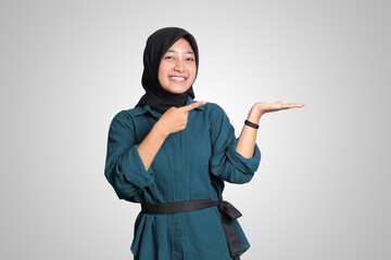 Portrait of excited Asian hijab woman in casual outfit standing against white background, showing product and pointing with hand and finger to the side. Advertising concept.