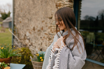 Profile portrait of lovely cute lady with dark hair looking down, holding plaid on shoulders and resting on summer terrace near the house 