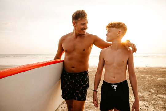 Smiling man holding paddleboard with arm around son at beach during sunset - Powered by Adobe