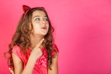 Happy smiling little girl in pink dress pointing index finger up at pink background, looking away. Funny blonde lady expressing emotion, showing. Childhood emotional concept. Copy advertising space