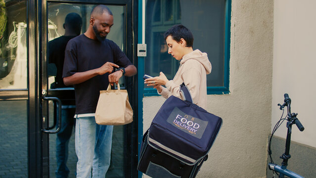 Male customer taking restaurant food order using smartwatch to pay with nfc on pos, paying for takeaway lunch. Food delivery service courier giving fastfood in paperbag, contactless terminal.