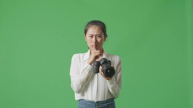 Asian Photographer Using A Camera Taking Pictures And Making Shh Gesture While Standing On Green Screen Background In The Studio
