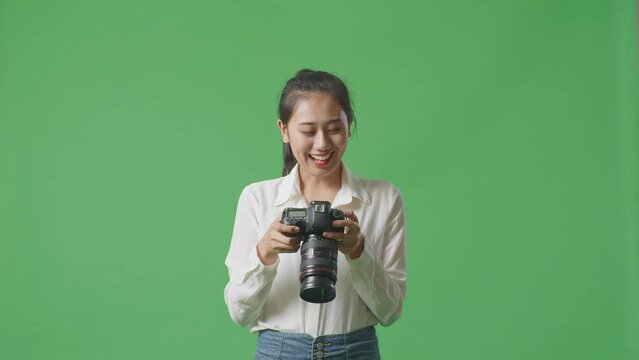 Asian Photographer Looking At The Pictures In The Camera And Smiling Being Satisfied With The Result While Standing On Green Screen Background In The Studio
