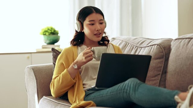 Coffee, laptop and Asian woman on couch, headphones and relax in home, calm and watching video. Japan, female and happy lady with headset, device and tea in living room, happiness and social media