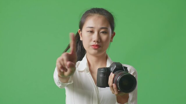Close Up Of Serious Asian Photographer Holding A Camera In Her Hands And Disapproving With No Index Finger Sign While Standing On Green Screen Background In The Studio
