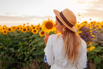 Back view of woman enjoying view in blooming sunflower field at sunset with bouquet of flowers....