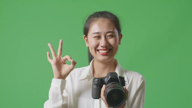 Close Up Of Asian Photographer Looking At The Pictures In The Camera And Showing Okay Gesture While Standing On Green Screen Background In The Studio
