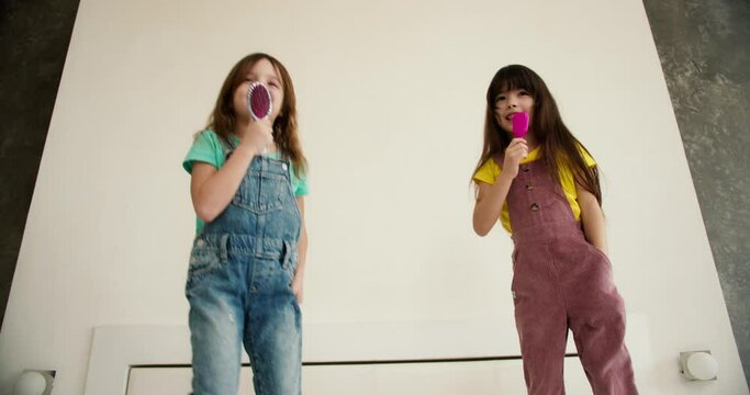 Funny cute little girls dancing singing with comb and have fun on bed at home, low angle view