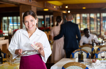 Smiling young female waitress standing with tray with tip in restaurant