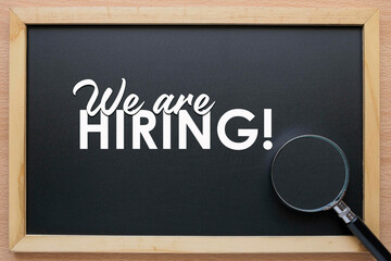 A black chalkboard with the message on We Are Hiring and a magnifying glass at the side. Human resource conept