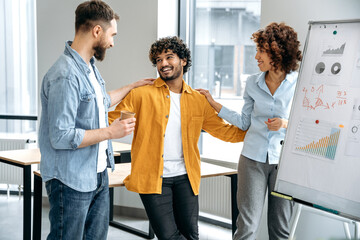 Motivated group of multiracial colleagues, creative team of top managers, stand in a modern office near a white marker board, communicate, are satisfied with the results of work, smile