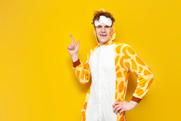 young joyful guy in funny baby giraffe pajamas and sleep mask shows his hand to the side on yellow...