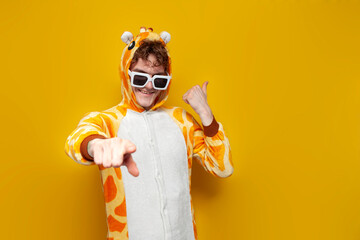 young joyful guy in funny children's giraffe pajamas and glasses invites you to party on yellow...