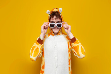 young joyful guy in funny baby giraffe pajamas and glasses on yellow background, man in animal...