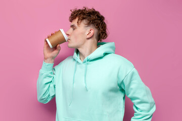 curly guy in mint hoodie drinks coffee from paper cup on pink isolated background, man in blue...
