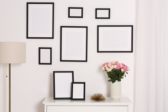 Empty frames hanging on white wall and chest of drawers with flowers indoors