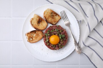 Tasty beef steak tartare served with yolk, capers, toasted bread and greens on white tiled table, top view