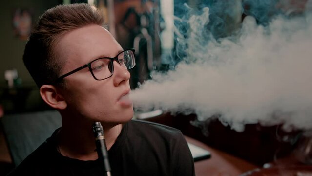 A hookah man in glasses smokes a traditional hookah pipe A man exhales smoke in a hookah cafe or lounge bar close-up