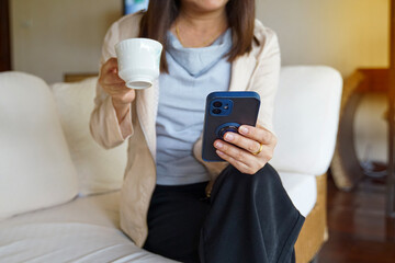Fototapeta premium Asian woman sitting on sofa sipping coffee and watching online news on mobile phone in the morning before going to work. Soft and selective focus. 