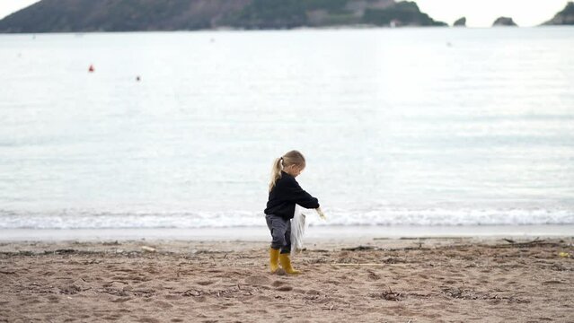 Little girl in gloves collects waste in a bag on the seashore